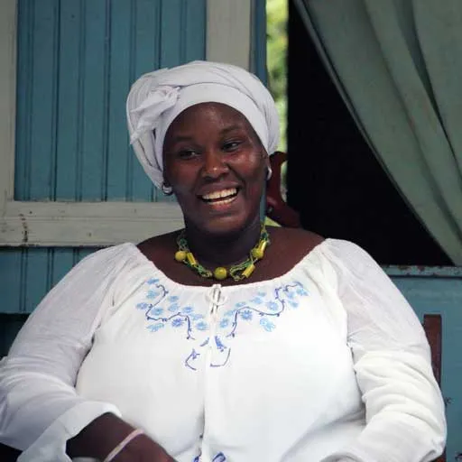 Retrato de una mujer sonriente con turbante blanco y blusa blanca bordada, sentada frente a una pared de madera azul.