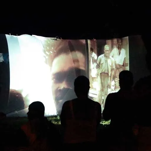Proyección nocturna de fotografías antiguas sobre una pantalla al aire libre, con siluetas de público en primer plano.