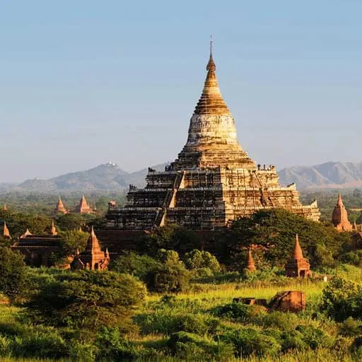 Templo de piedra escalonado de Bagan con varias estupas menores entre vegetación, con montañas al fondo.